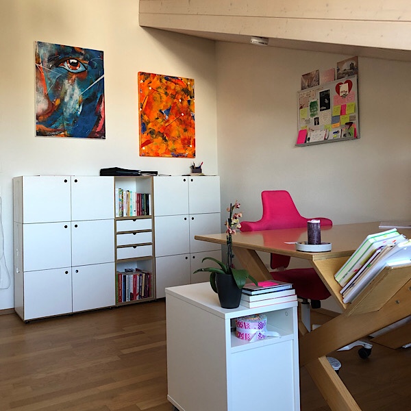 Schreibtisch mit pinkem Stuhl, weissem Sideboard und Bildern im Hintergrund 