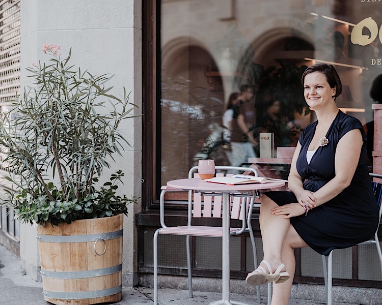 Christine in blauem Kleid und weissen Sandalen vor einem Café mit Olivenbaum